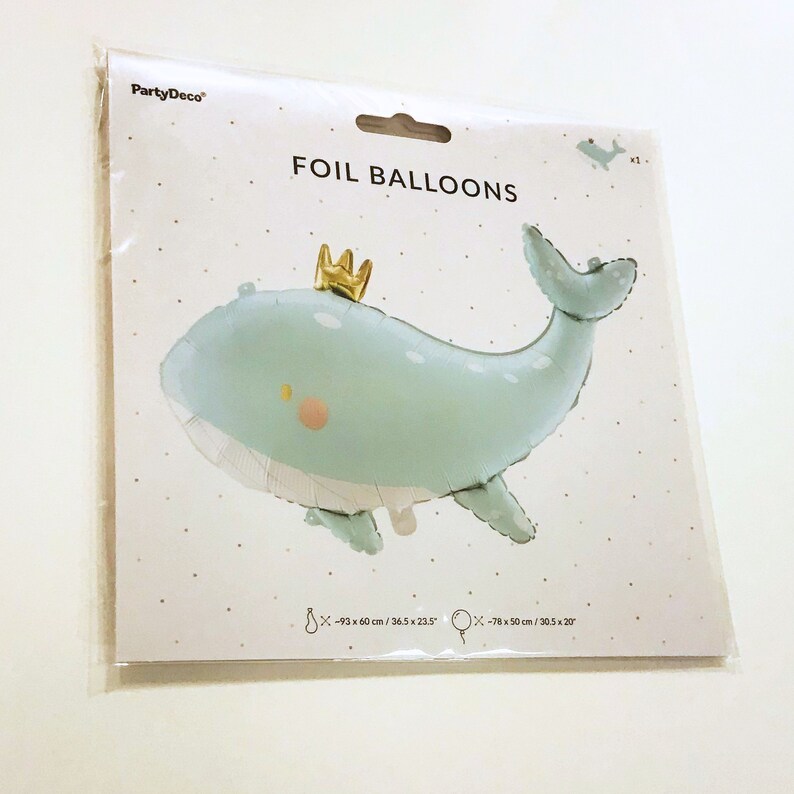 Może przedstawiać: Jasnoniebieski balon foliowy w kształcie wieloryba z złotą koroną na głowie. Balon ma białe kropki i r&oacute;żowy rumieniec na policzku. Opakowanie zawiera napis "FOIL BALLOONS" i podaje wymiary balonu w calach i centymetrach.