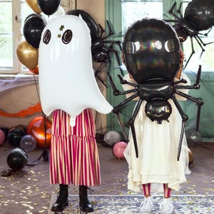 May include: Two children holding Halloween-themed balloons. One child holds a white ghost balloon with black eyes and star accents. The other holds a black spider balloon. The background includes other balloons and Halloween decorations.
