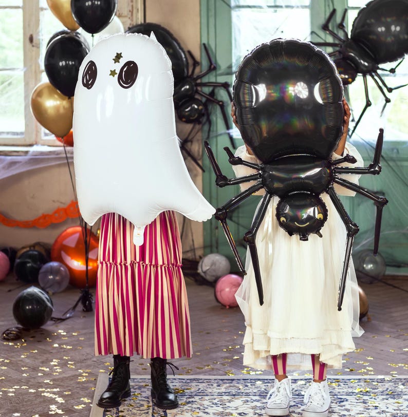 May include: Two children holding Halloween-themed balloons. One child holds a white ghost balloon with black eyes and star accents. The other holds a black spider balloon. The background includes other balloons and Halloween decorations.