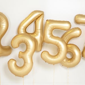 May include: Gold foil number balloons, including the numbers 1, 2, 3, 4, 5, 6, 7, and 8. The balloons are inflated and tied together.