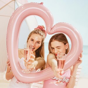 Two women posing inside a large pink heart balloon frame at the beach, fun bachelorette party and bridal shower photo decoration