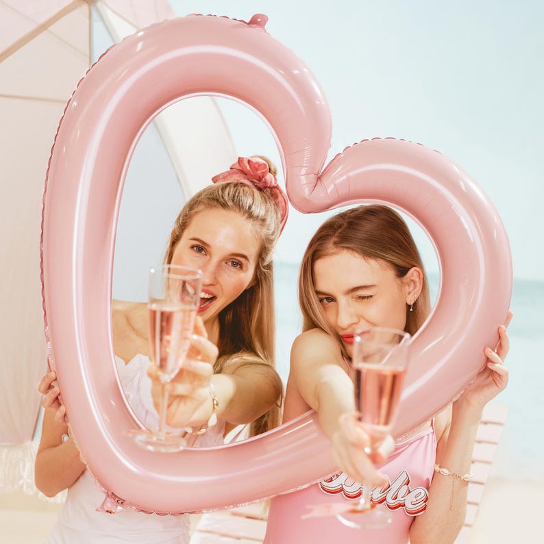 Two women posing inside a large pink heart balloon frame at the beach, fun bachelorette party and bridal shower photo decoration