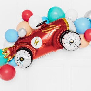 May include: A red race car shaped balloon with a gold lightning bolt and white wheels surrounded by a variety of colorful balloons.