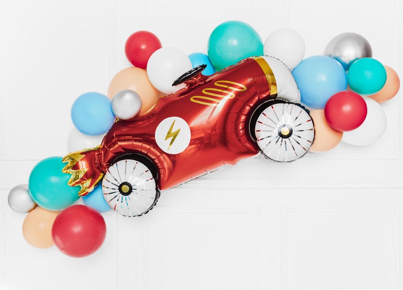 May include: A red race car shaped balloon with a gold lightning bolt and white wheels surrounded by a variety of colorful balloons.