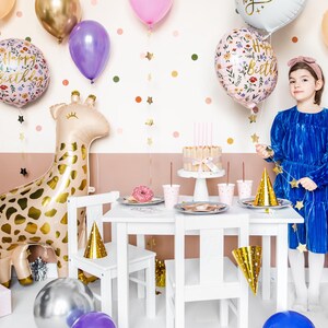 May include: A birthday party table setting with a white table, gold star garland, and a giraffe balloon. There are pink and blue cups, a cake, and a gold party hat on the table. A girl in a blue dress is holding a gold star garland. The text on the balloon says "Happy Birthday".