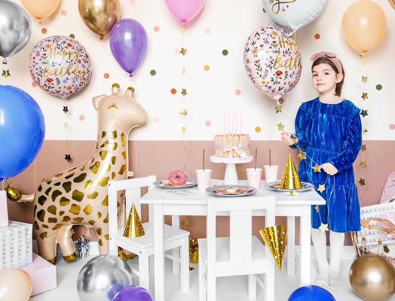 May include: A birthday party table setting with a white table, gold star garland, and a giraffe balloon. There are pink and blue cups, a cake, and a gold party hat on the table. A girl in a blue dress is holding a gold star garland. The text on the balloon says "Happy Birthday".