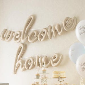 Op de afbeelding: Een beige ballonslinger met de tekst "welcome home" hangt aan een muur. Witte, lichtblauwe en roze ballonnen met de tekst "Welcome Baby" zijn rechts gegroepeerd. Een taart en cupcakes staan op een tafel eronder.