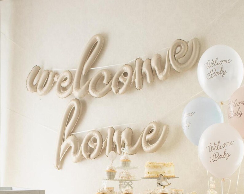 Op de afbeelding: Een beige ballonslinger met de tekst "welcome home" hangt aan een muur. Witte, lichtblauwe en roze ballonnen met de tekst "Welcome Baby" zijn rechts gegroepeerd. Een taart en cupcakes staan op een tafel eronder.