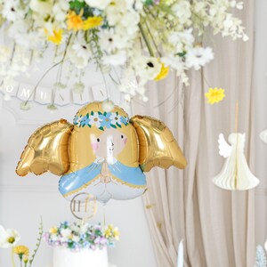 以下が含まれることがあります： 白い天使のデザインと青いドレスが付いた金のホイルバルーン。バルーンは翼と花冠が付いています。バルーンは白い天井から吊り下げられており、白い文字で「KOMUNIA」と書かれたバナーが付いています。