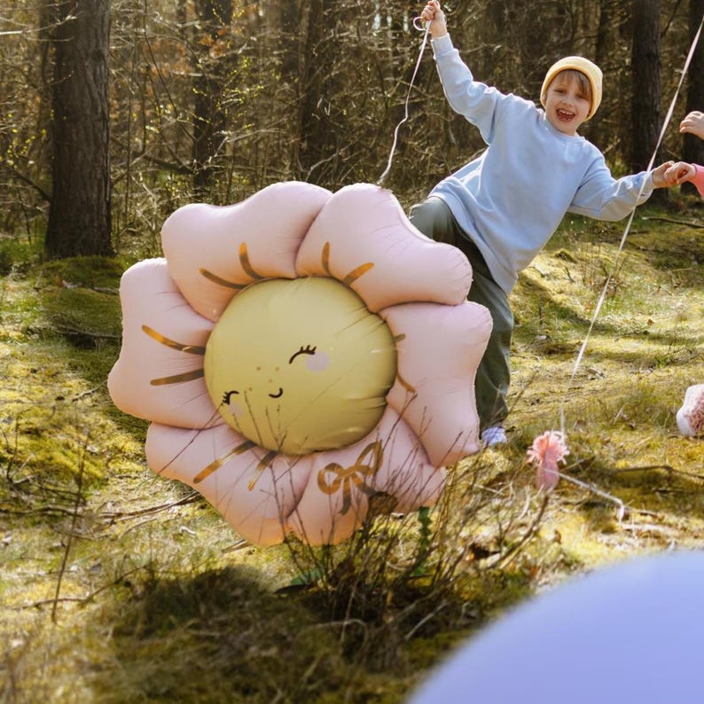 May include: A large pink and yellow inflatable flower balloon with a smiling face. The balloon is being held by a child in a blue sweater and a yellow hat in a forest setting.