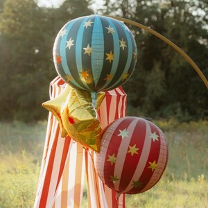 K&ouml;nnte beinhalten: Eine Sammlung festlicher Ballons auf einem Feld. Die Ballons umfassen eine gro&szlig;e, blaugr&uuml;n und schwarz gestreifte Kugel mit goldenen Sternen, einen goldfarbenen sternf&ouml;rmigen Ballon und eine rot-wei&szlig; gestreifte Kugel mit goldenen Sternen. Im Hintergrund befindet sich ein rot-wei&szlig; gestreiftes Zelt.