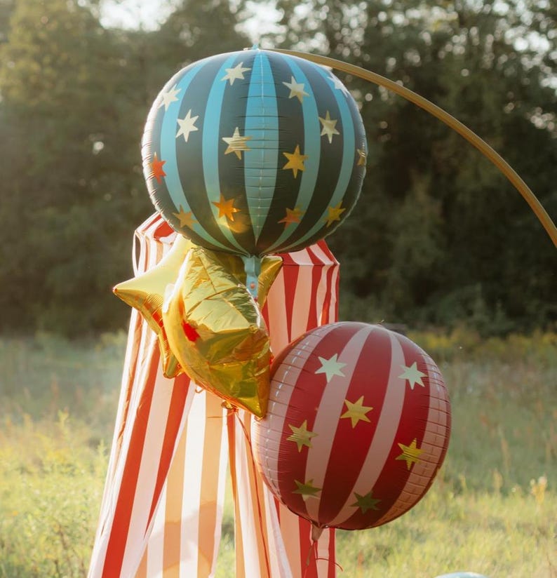 K&ouml;nnte beinhalten: Eine Sammlung festlicher Ballons auf einem Feld. Die Ballons umfassen eine gro&szlig;e, blaugr&uuml;n und schwarz gestreifte Kugel mit goldenen Sternen, einen goldfarbenen sternf&ouml;rmigen Ballon und eine rot-wei&szlig; gestreifte Kugel mit goldenen Sternen. Im Hintergrund befindet sich ein rot-wei&szlig; gestreiftes Zelt.