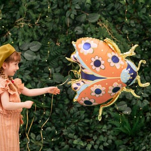 May include: A young girl wearing a brown beret and a peach-colored dress holds a string attached to a large orange, blue, and white ladybug balloon. The balloon has a floral pattern and is against a green leafy background.
