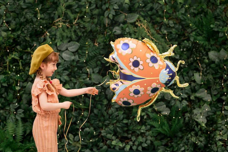 May include: A young girl wearing a brown beret and a peach-colored dress holds a string attached to a large orange, blue, and white ladybug balloon. The balloon has a floral pattern and is against a green leafy background.