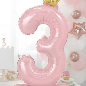 Puede incluir: Un globo de n&uacute;mero tres de color rosa con una corona dorada en la parte superior. El globo est&aacute; de pie sobre una base rosa.