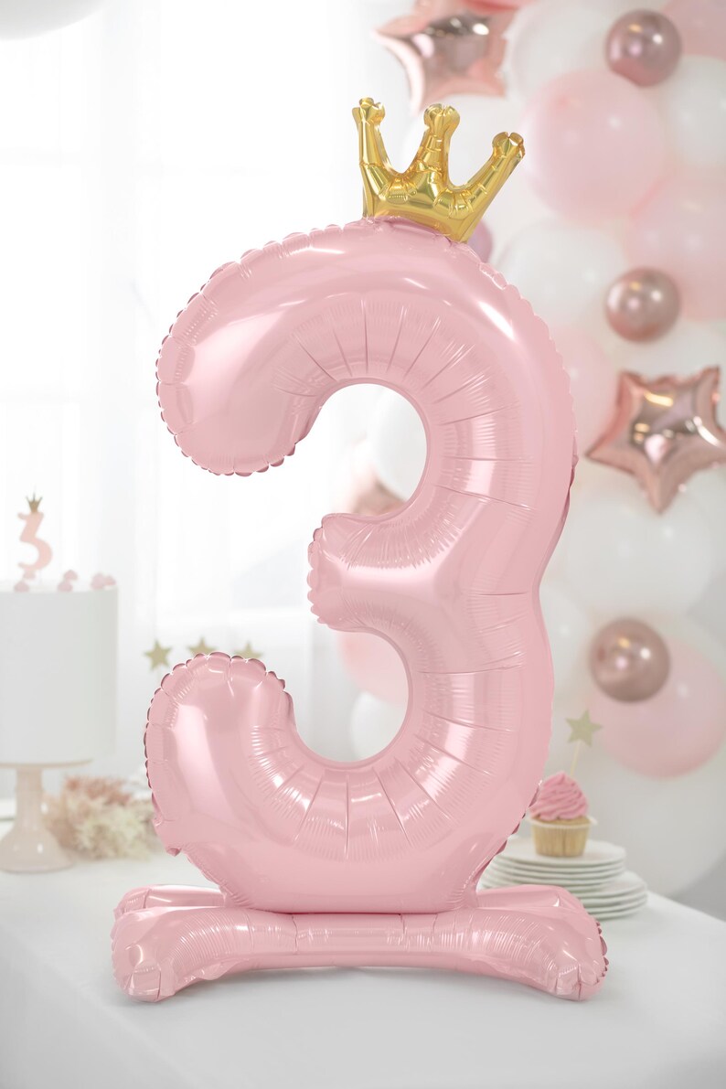 Puede incluir: Un globo de n&uacute;mero tres de color rosa con una corona dorada en la parte superior. El globo est&aacute; de pie sobre una base rosa.