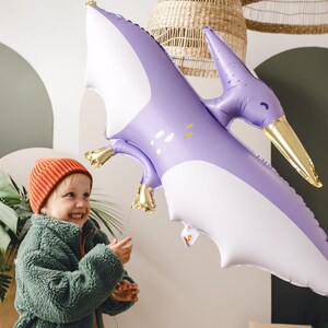 May include: A child wearing an orange beanie and a green jacket smiles while holding a purple and white inflatable pterodactyl balloon.