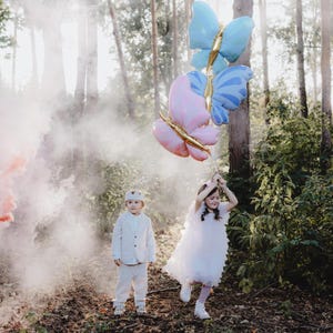 Pu&ograve; includere: Due bambini, un ragazzo e una ragazza, stanno giocando in un bosco con palloncini a forma di farfalla colorati. La ragazza indossa un vestito bianco e il ragazzo indossa un abito bianco. I palloncini sono rosa e blu e hanno dettagli dorati.