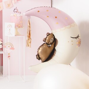 Pu&ograve; includere: Un palloncino a forma di mezzaluna rosa e bianco con accenti di glitter dorati e un piccolo palloncino di orso marrone all'interno. Il palloncino a forma di luna ha un fiocco dorato che pende da esso. Il testo "Oh Baby" &egrave; visibile sullo sfondo.