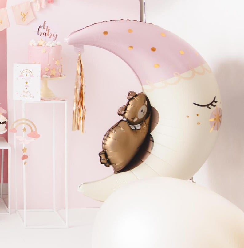 Pu&ograve; includere: Un palloncino a forma di mezzaluna rosa e bianco con accenti di glitter dorati e un piccolo palloncino di orso marrone all'interno. Il palloncino a forma di luna ha un fiocco dorato che pende da esso. Il testo "Oh Baby" &egrave; visibile sullo sfondo.