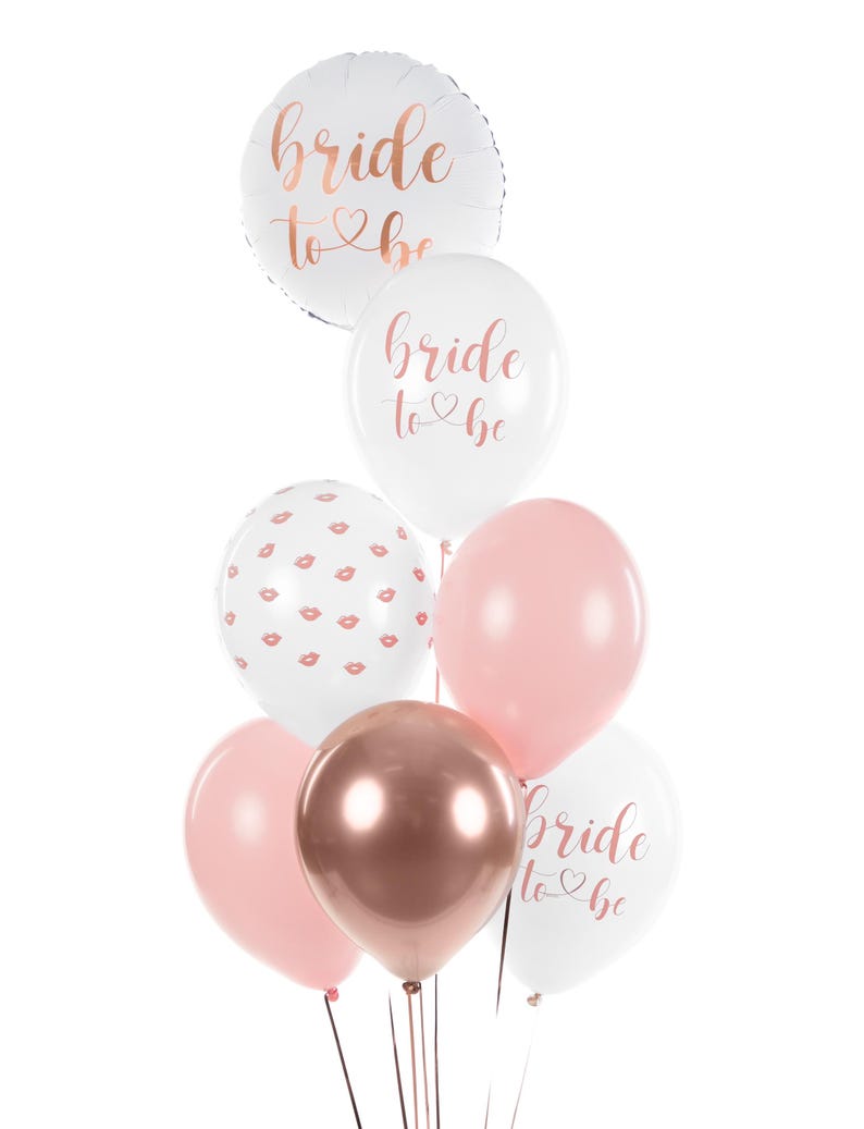 May include: A bouquet of balloons for a bridal shower or bachelorette party. The balloons are white, pink, and rose gold. Some of the balloons have the text "bride to be" printed on them in rose gold lettering. One balloon has a pattern of pink lips on a white background.