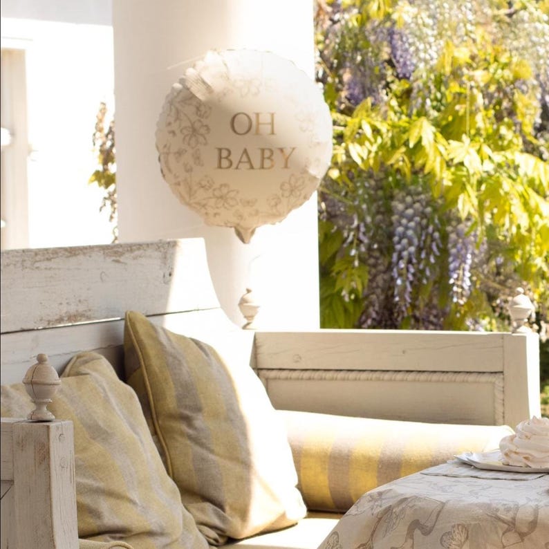 Puede incluir: Un globo redondo color crema con el texto dorado "OH BABY". El globo est&aacute; decorado con un patr&oacute;n floral y cuelga frente a un banco blanco con cojines a rayas amarillas. La escena es al aire libre, con vegetaci&oacute;n de fondo.