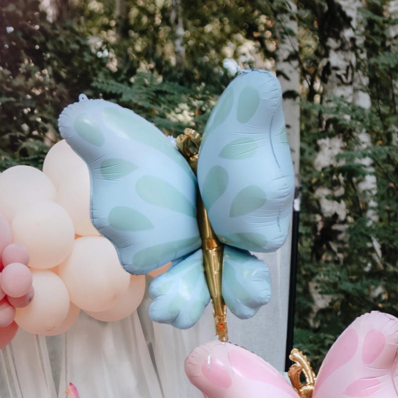 Pu&ograve; includere: Un palloncino a forma di farfalla blu chiaro con un centro dorato. Il palloncino &egrave; decorato con un motivo verde chiaro. Il palloncino &egrave; circondato da palloncini bianchi e rosa.