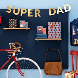 May include: A festive Father's Day scene with a red bicycle, gifts, and balloons. A banner reads "SUPER DAD" in gold letters. A blue #1 Dad foam finger balloon and a round balloon with "Super Dad" are also featured.