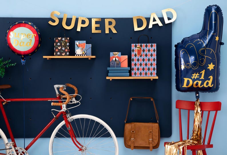 May include: A festive Father's Day scene with a red bicycle, gifts, and balloons. A banner reads "SUPER DAD" in gold letters. A blue #1 Dad foam finger balloon and a round balloon with "Super Dad" are also featured.