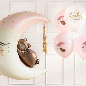 May include: A pink and white crescent moon balloon with a brown bear sleeping inside. The balloon is surrounded by smaller pink and white balloons with a bear and moon design. The balloons have the text "Baby girl" printed on them.