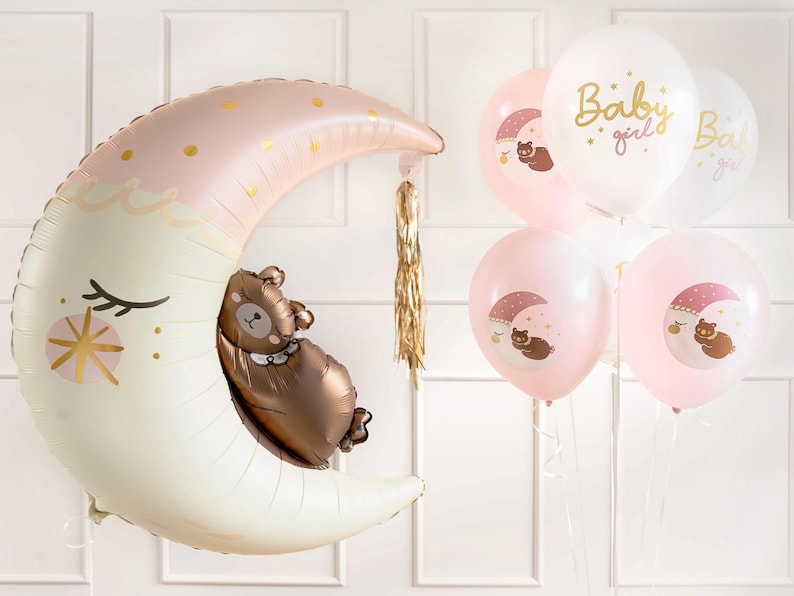 May include: A pink and white crescent moon balloon with a brown bear sleeping inside. The balloon is surrounded by smaller pink and white balloons with a bear and moon design. The balloons have the text "Baby girl" printed on them.