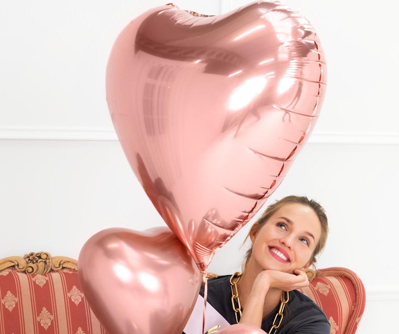 May include: A woman smiles while holding two large rose gold heart-shaped balloons.