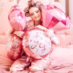 May include: A woman in pink pajamas holds a variety of pink and white balloons. The balloons have a diamond pattern and one says "She said yes".