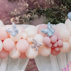 May include: A backdrop of white and pink balloons with blue butterfly decorations. The balloons are arranged in a cascading pattern, creating a festive and whimsical atmosphere.