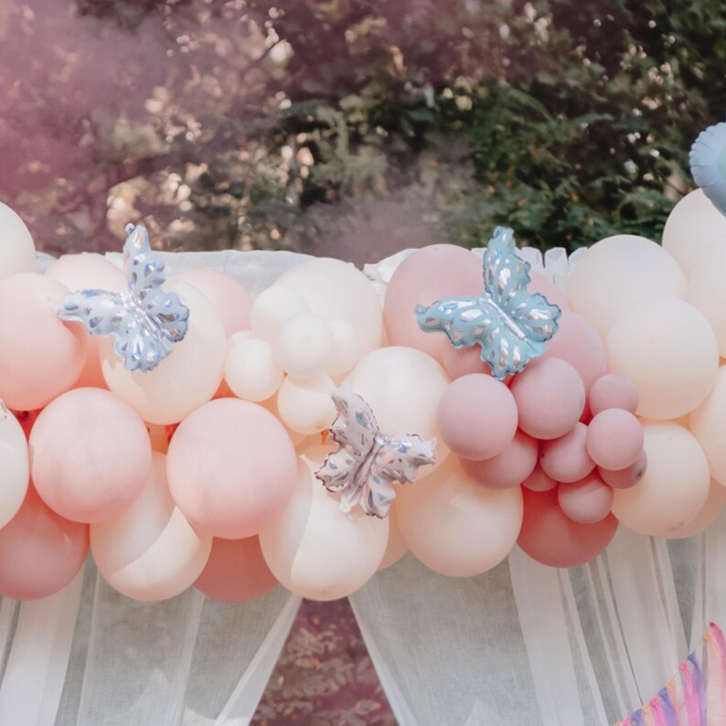 May include: A backdrop of white and pink balloons with blue butterfly decorations. The balloons are arranged in a cascading pattern, creating a festive and whimsical atmosphere.