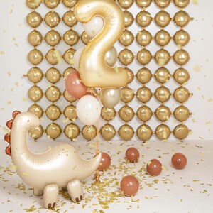 Celebration setup featuring a cream-colored dinosaur-shaped foil balloon with copper spikes, surrounded by gold, white, and copper balloons, including a large gold number "2" balloon. Background features a wall of small, round gold balloons arranged in a grid pattern, with gold confetti scattered on the floor.