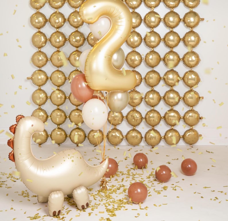 Celebration setup featuring a cream-colored dinosaur-shaped foil balloon with copper spikes, surrounded by gold, white, and copper balloons, including a large gold number "2" balloon. Background features a wall of small, round gold balloons arranged in a grid pattern, with gold confetti scattered on the floor.