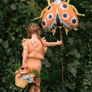 May include: A young person wearing a peach-colored jumpsuit with floral temporary tattoos on their back, holding a basket of flowers and a large orange and blue ladybug balloon in front of a green leafy wall.