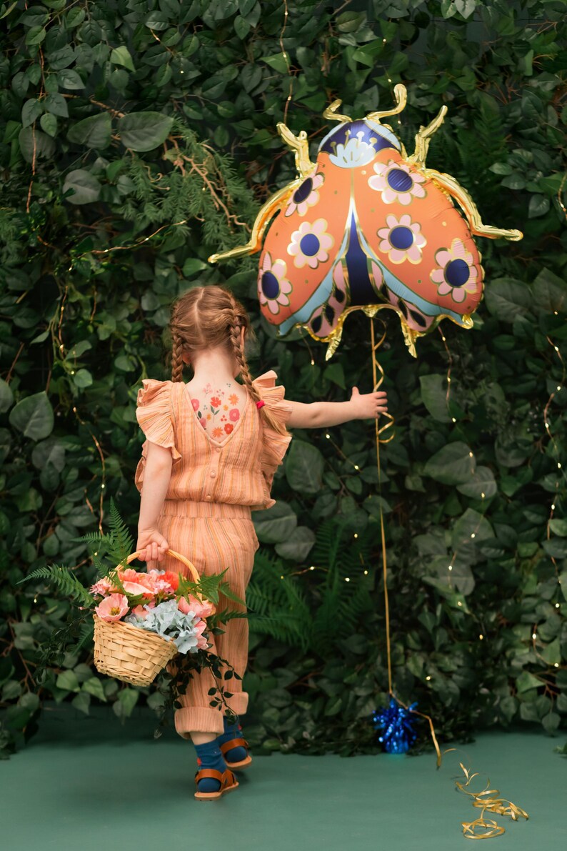 May include: A young person wearing a peach-colored jumpsuit with floral temporary tattoos on their back, holding a basket of flowers and a large orange and blue ladybug balloon in front of a green leafy wall.