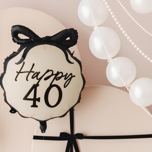 A festive scene featuring a cream-colored and black balloon with a bow design and the text "Happy 40" in elegant black lettering. The background includes white balloons and pearl-like garlands, along with large, round, cream-colored props tied with black ribbon, creating a sophisticated 40th celebration setup.