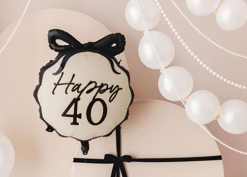 A festive scene featuring a cream-colored and black balloon with a bow design and the text "Happy 40" in elegant black lettering. The background includes white balloons and pearl-like garlands, along with large, round, cream-colored props tied with black ribbon, creating a sophisticated 40th celebration setup.