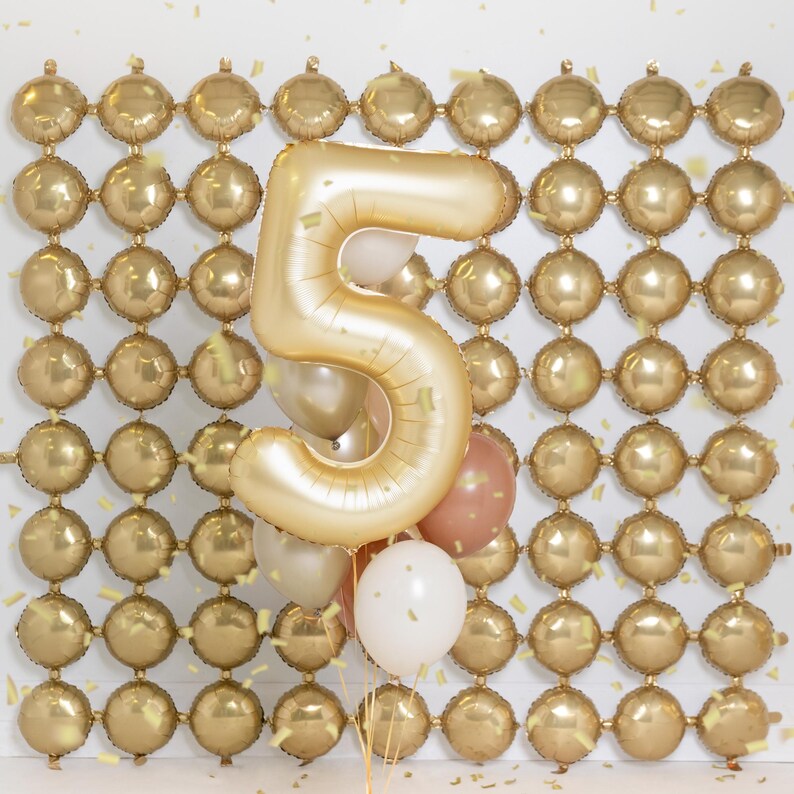 May include: A gold number five balloon with a cluster of smaller balloons in gold, white, and peach. The balloons are in front of a wall of gold balloons.