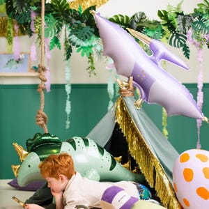 May include: A child is reading a book while sitting in a teepee decorated with green and purple dinosaur balloons, gold fringe, and green leaves. The teepee is on a pink and white rug with a dinosaur toy on the floor.