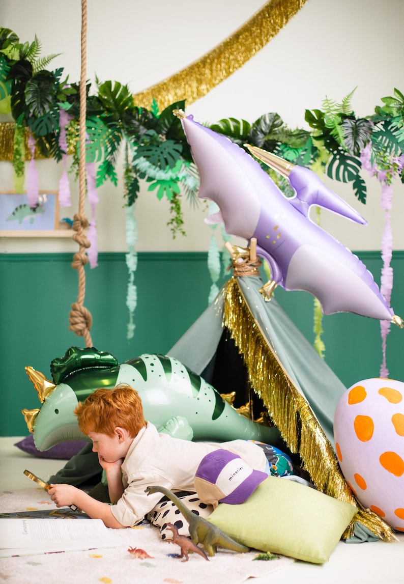 May include: A child is reading a book while sitting in a teepee decorated with green and purple dinosaur balloons, gold fringe, and green leaves. The teepee is on a pink and white rug with a dinosaur toy on the floor.