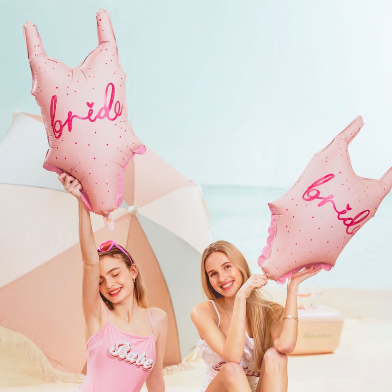 May include: Two women in pink swimsuits holding pink foil balloons shaped like a swimsuit with the word "bride" written on them. One woman is holding a pink umbrella.