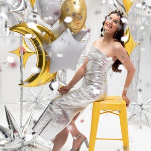 Op de afbeelding: Een vrouw in een zilveren jurk zit op een gele kruk, omringd door zilveren en gouden stervormige ballonnen en confetti. Ze houdt een gouden halvemaanballon vast.