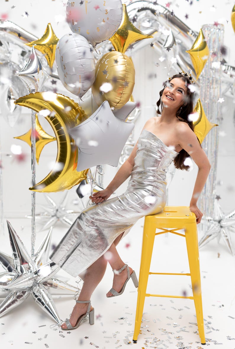 Op de afbeelding: Een vrouw in een zilveren jurk zit op een gele kruk, omringd door zilveren en gouden stervormige ballonnen en confetti. Ze houdt een gouden halvemaanballon vast.