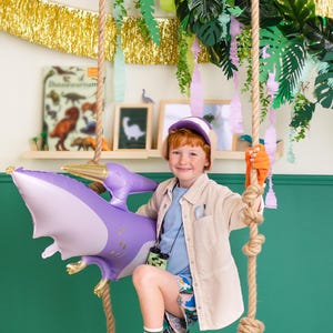 May include: A young boy dressed as a dinosaur explorer is standing on a large purple and orange dinosaur egg. He is holding a purple inflatable pterodactyl balloon and wearing a brown safari jacket, blue shirt, and a dinosaur hat.
