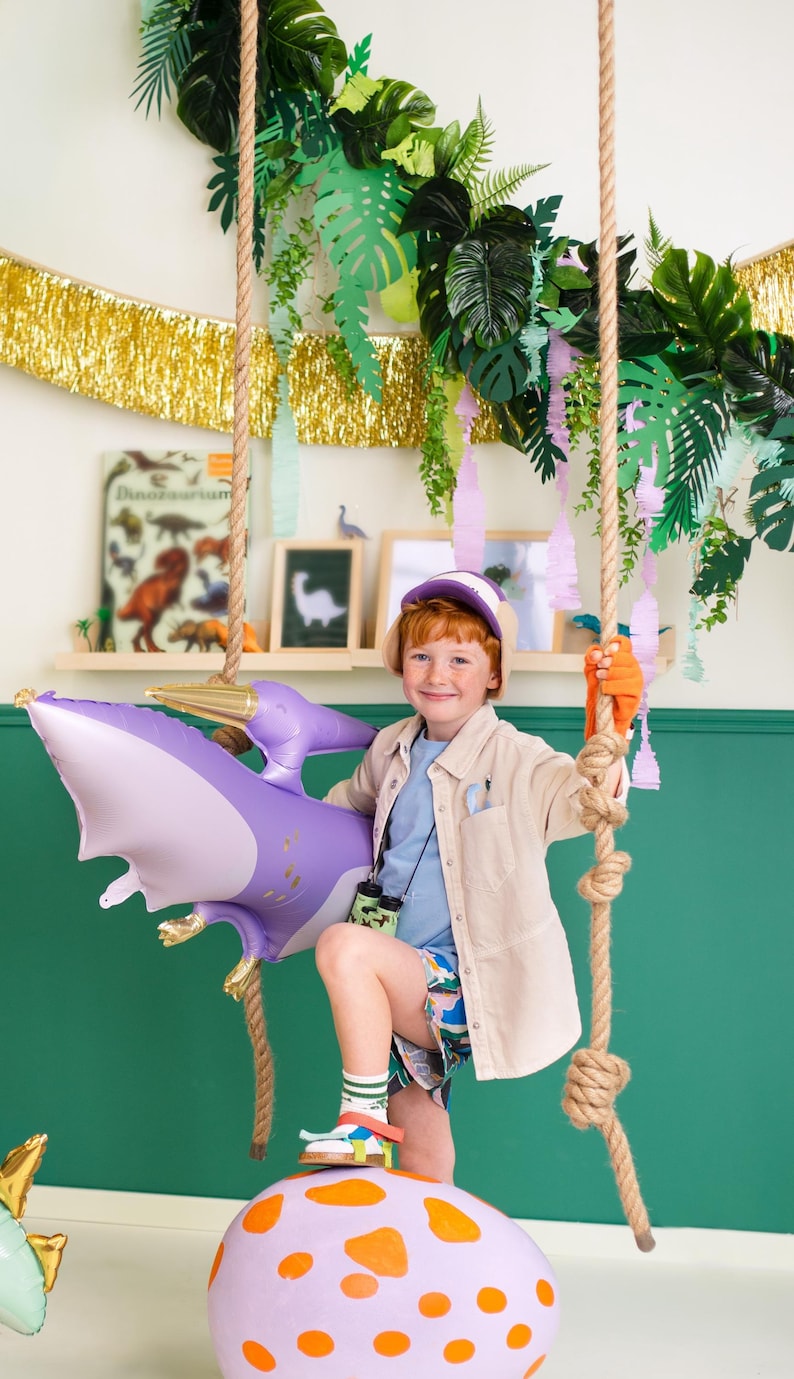 May include: A young boy dressed as a dinosaur explorer is standing on a large purple and orange dinosaur egg. He is holding a purple inflatable pterodactyl balloon and wearing a brown safari jacket, blue shirt, and a dinosaur hat.