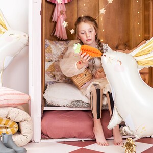 May include: A young girl sits in a white wooden cubby with a white and gold bunny balloon. She is holding a carrot-shaped balloon and a wicker basket. The cubby is decorated with pink and white pillows and blankets.